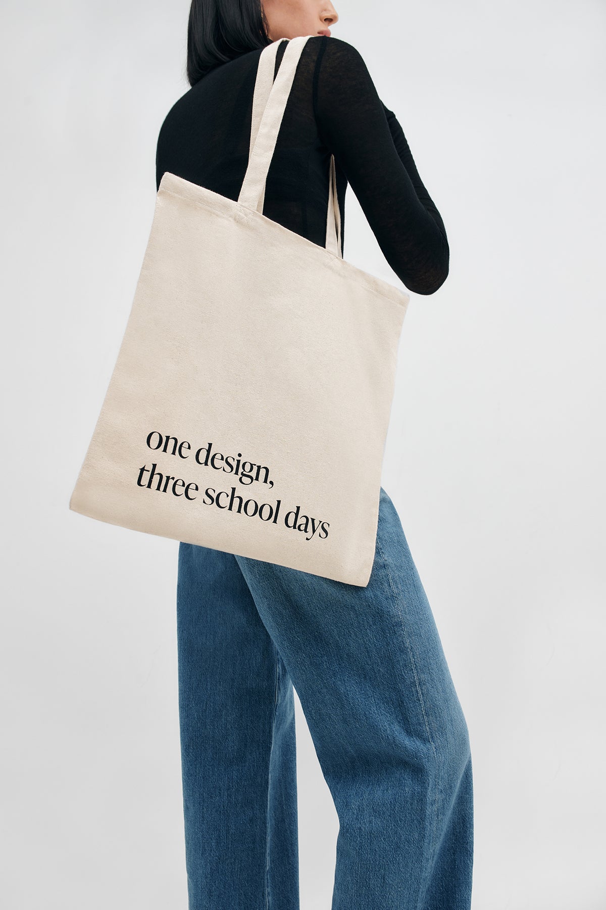 Cream canvas tote bag with black text reading "one design. three school days" worn over shoulder.