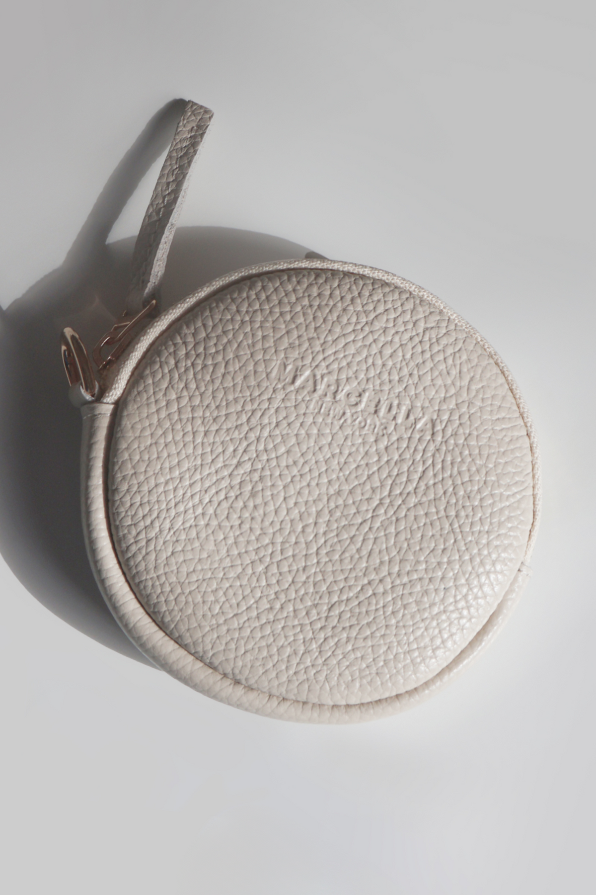 Cream circular pouch with textured leather surface and woven handle against neutral background.