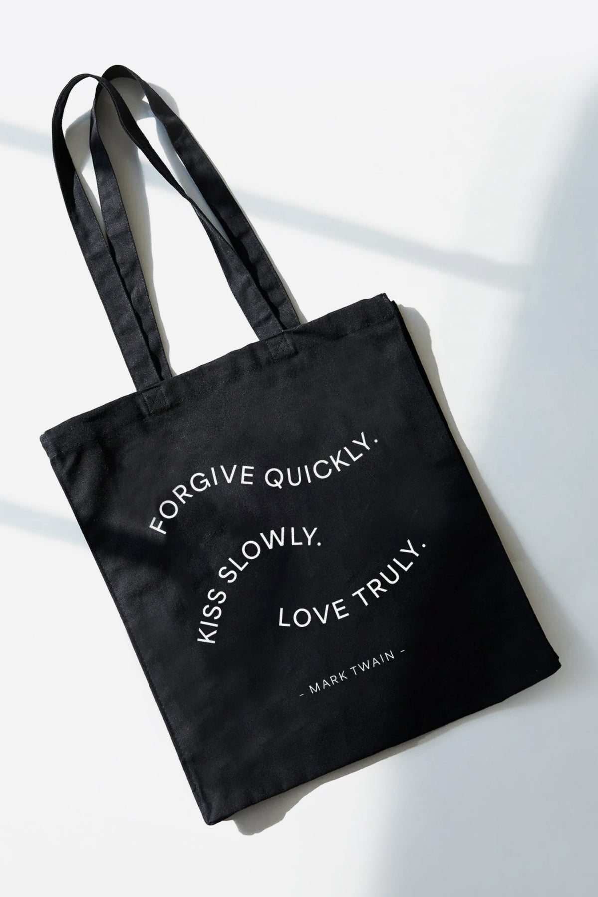 Black tote bag with white text reading "Forgive quickly. Kiss slowly. Love truly. - Mark Twain" and dual handles.