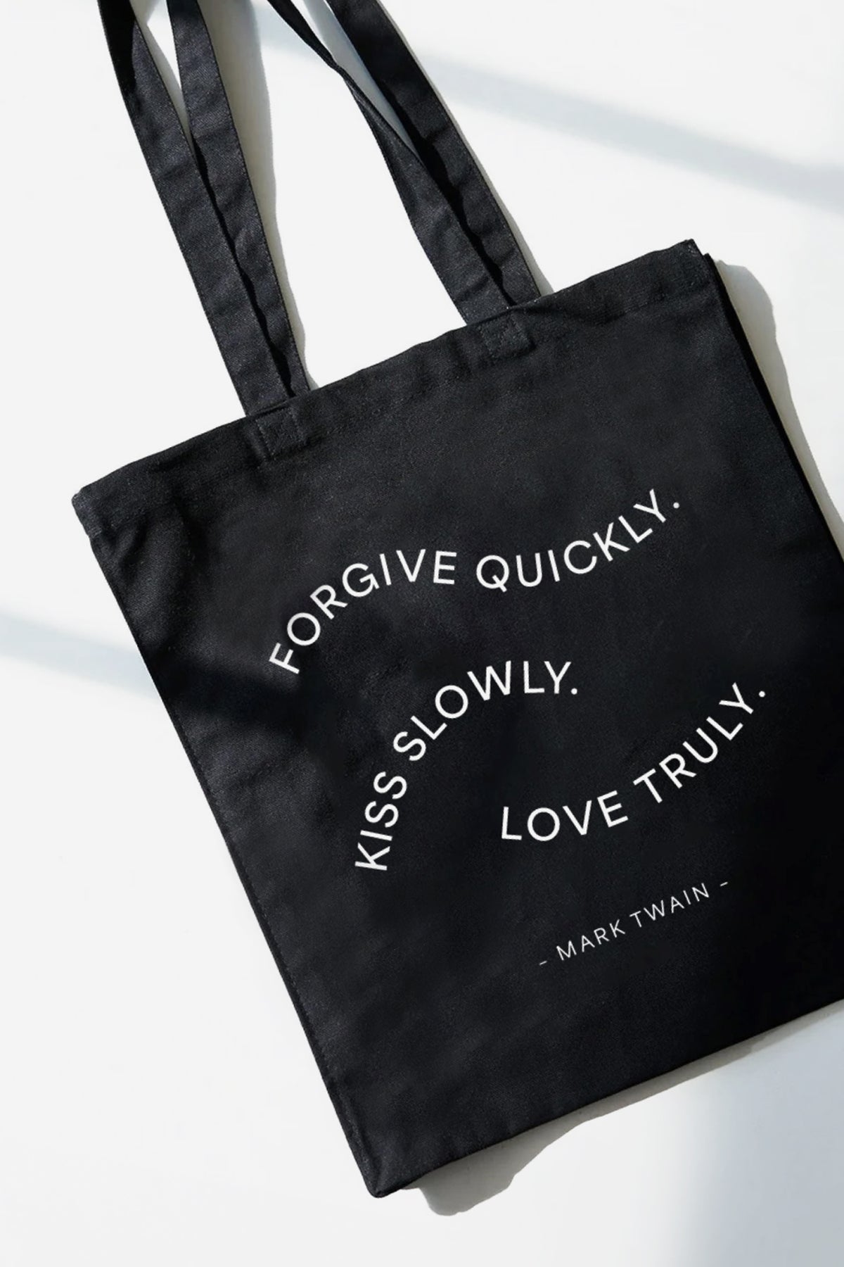 Black tote bag with white text reading "Forgive quickly. Kiss slowly. Love truly. - Mark Twain" and dual shoulder straps.