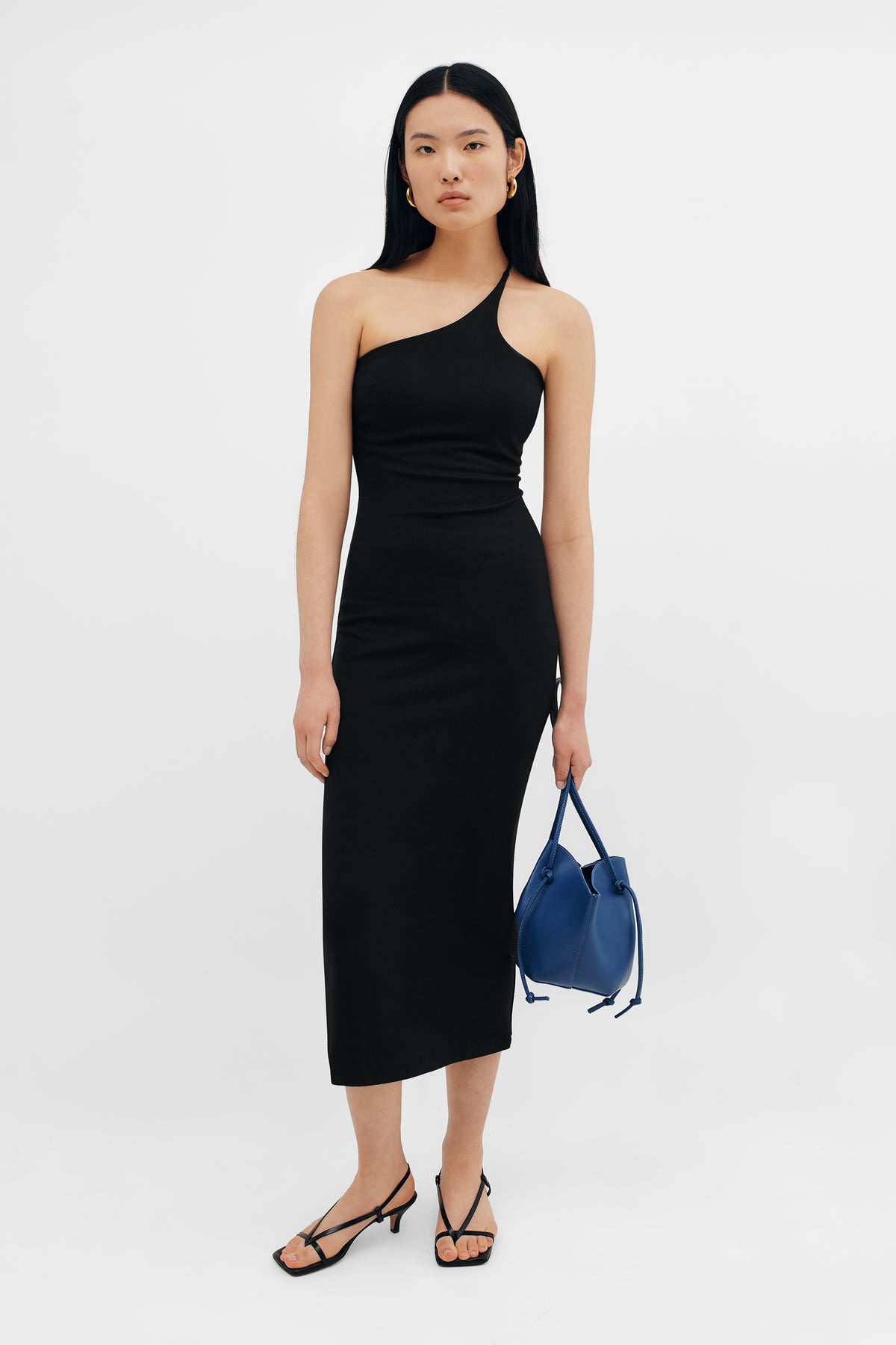Woman wearing black one-shoulder fitted midi dress with blue handbag and black strappy sandals.