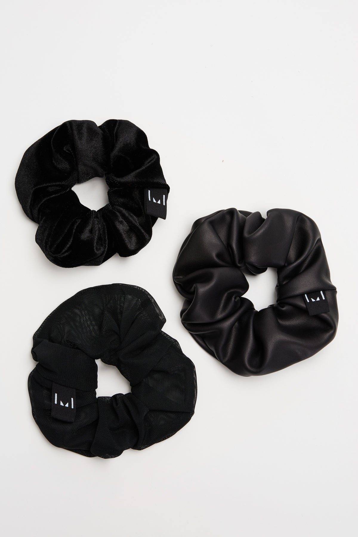 Three black scrunchies in velvet and textured fabrics with small branded tags arranged on white background.