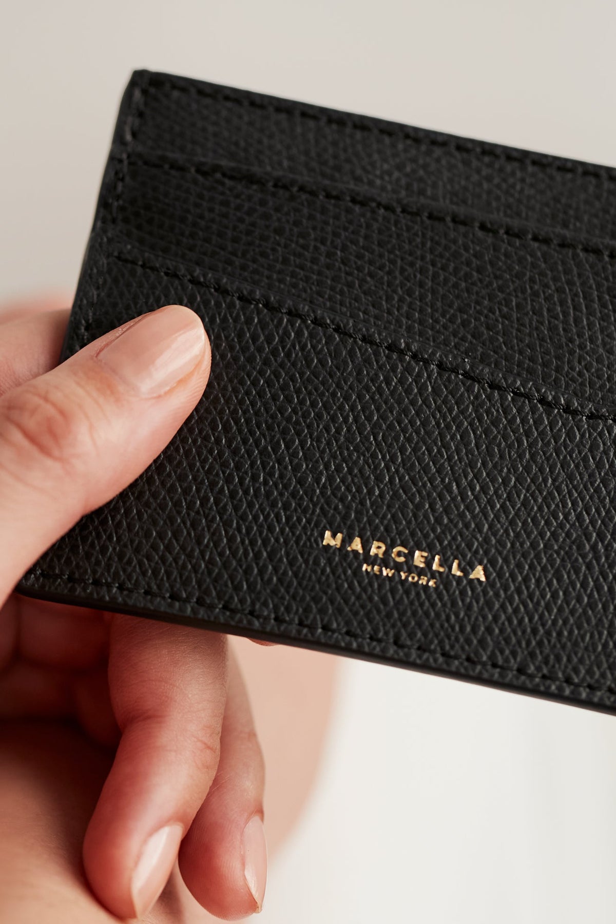 Black textured leather card holder with gold "Marcella New York" branding held in hand.