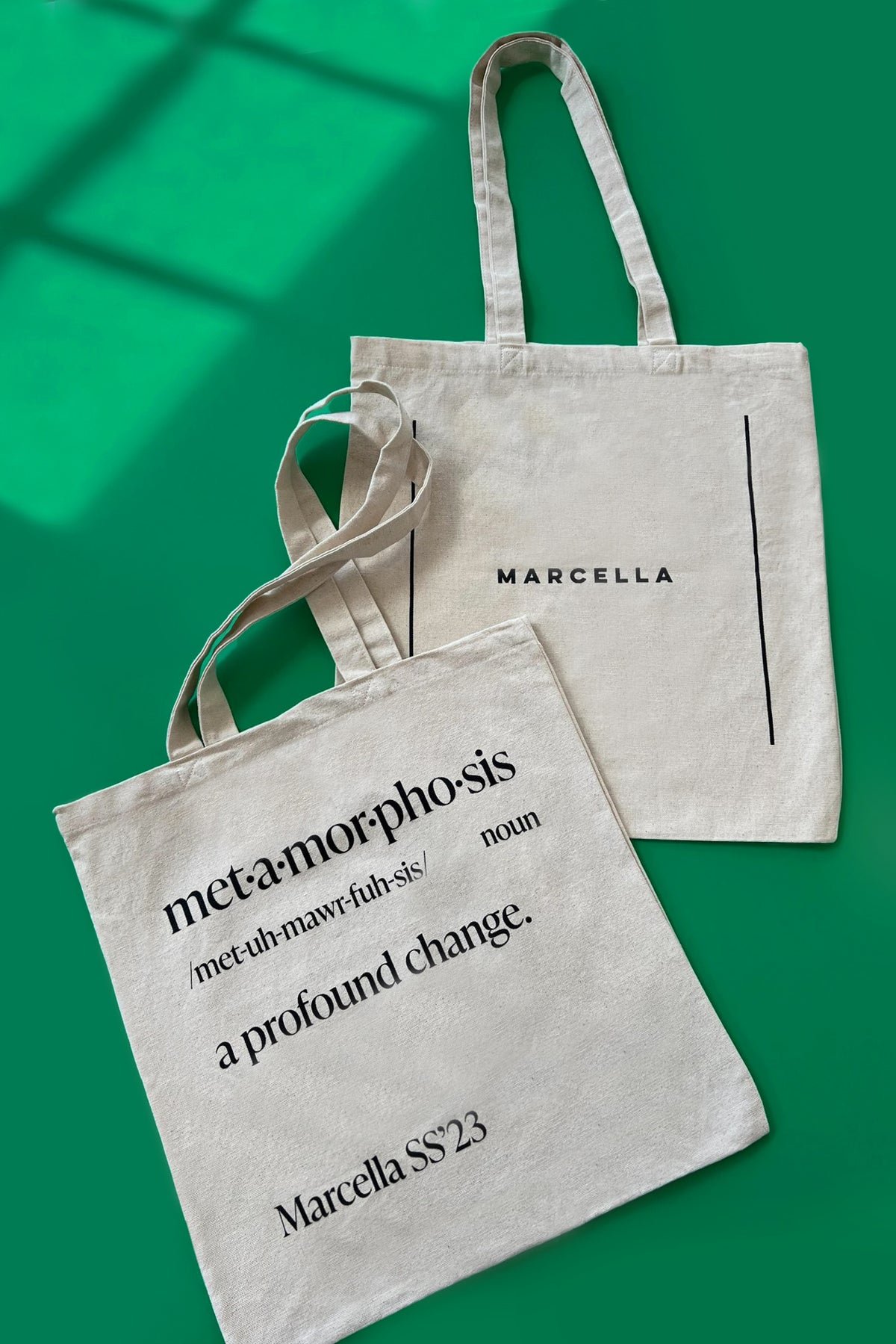 Beige cotton tote bags with "Marcella" branding and "metamorphosis" text definition printed on front, displayed against green