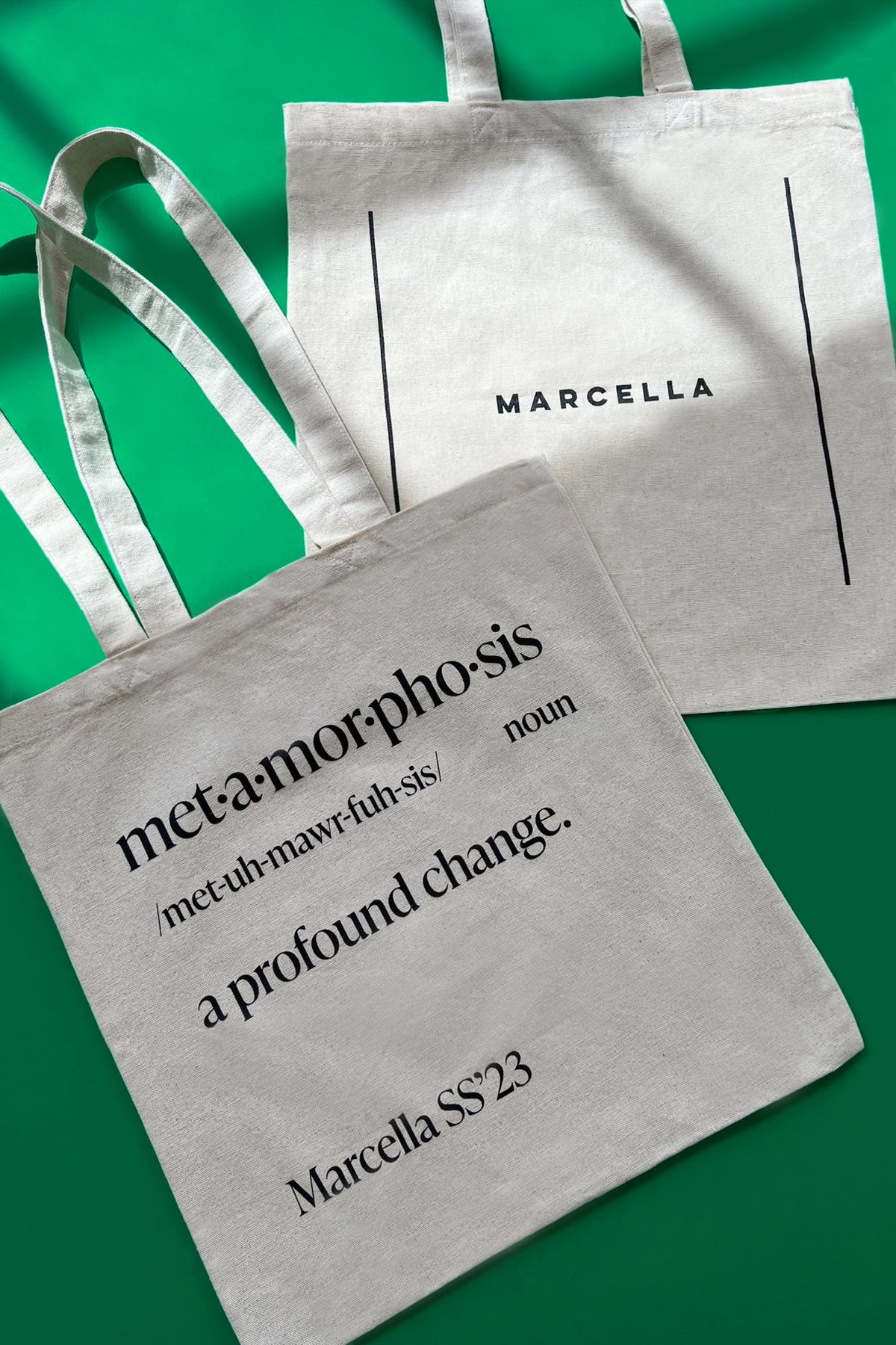 Cream canvas tote bags with black text reading "metamorphosis" definition and "Marcella" branding on green background.